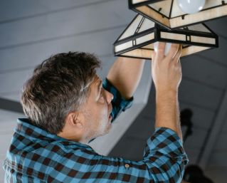 Adult male fixing pendant light indoors, wearing plaid long sleeves. Home maintenance scene.